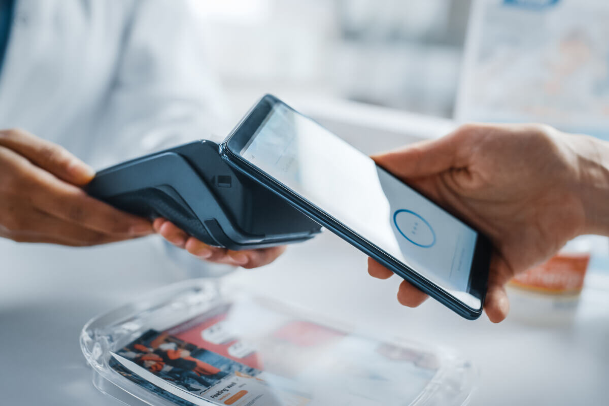 Close up of a contactless payment. One person holds their phone to a payment terminal that is held by a medical provider in a white coat.