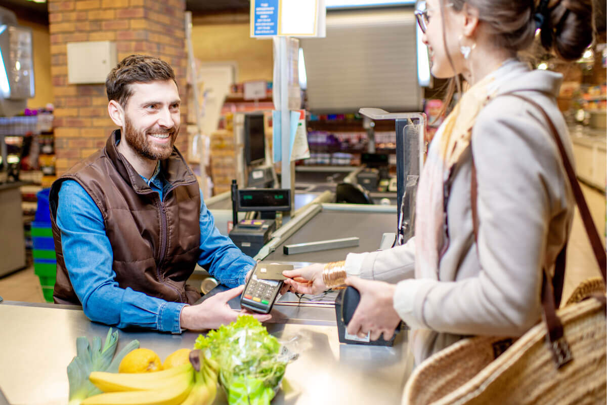 A woman checks out at a grocery store by holding her credit card against a handheld tap-to-pay payment terminal, which is held by a smiling male employee wearing a brown vest.