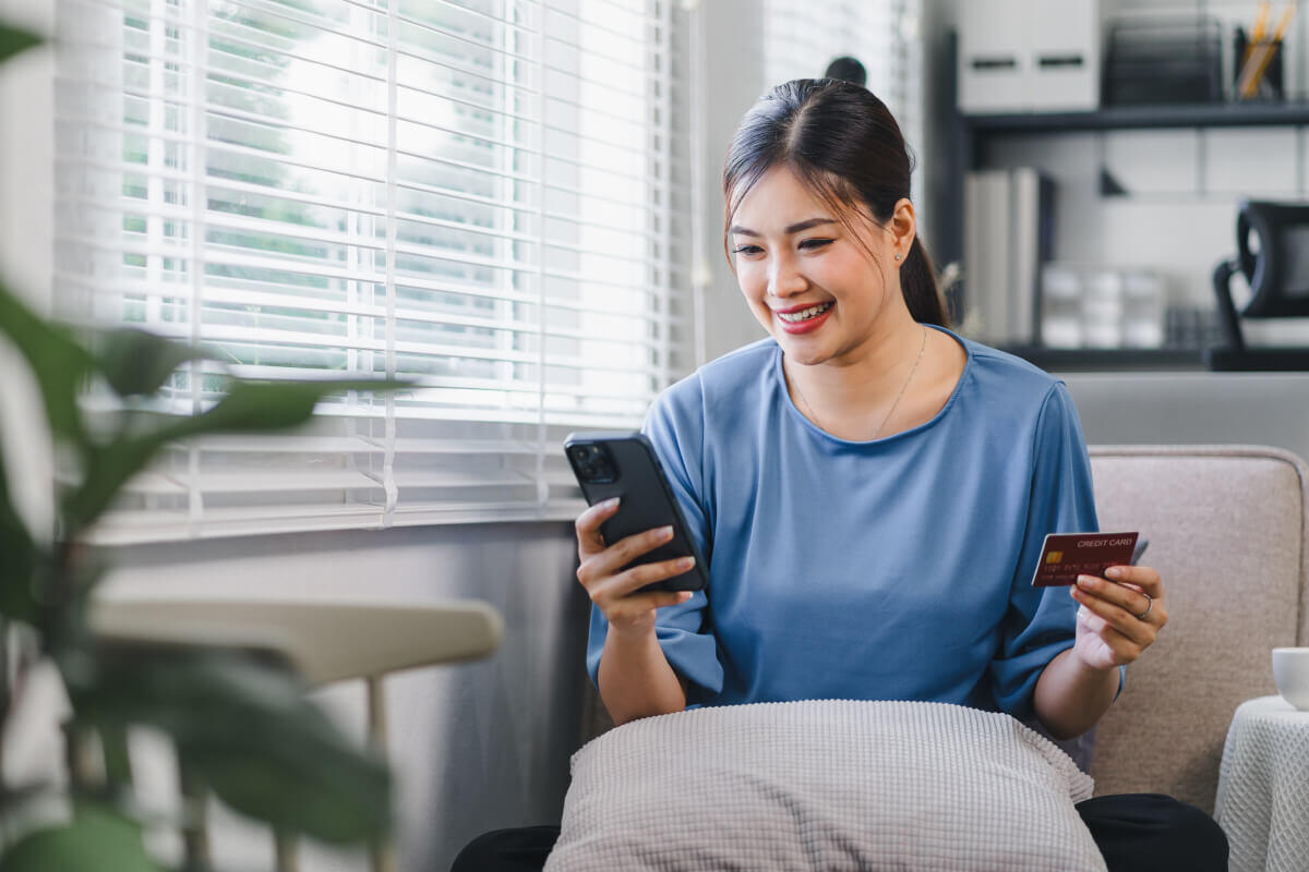 Young woman of Asian heritage sits in her home by a window. She holds her phone in one hand and her credit card in the other, completing a text to pay transaction.