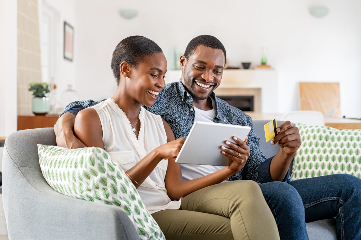 Black couple sitting on a gray couch. The woman is holding a silver tablet and pointing to something, while the man is holding a yellow credit card.