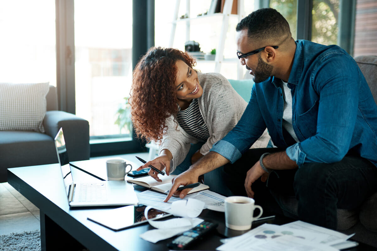 A Black couple sit at their living room couch and look at each other, smiling. There are documents, a laptop, and coffee mugs on the coffee table in front of them as they pay their bills.