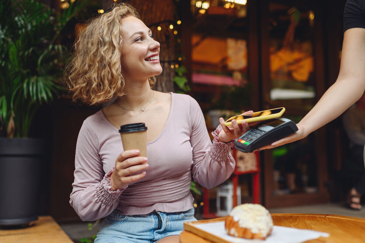 Woman with short blonde hair wearing a purple shirt completes a cashless payment. She holds a to-go coffee cup in one hand and her smartphone in the other, which is touching a handheld payment terminal, held by an employee out of frame.