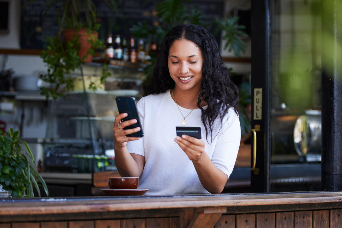 Woman with long black hair sits at the outside of a cafe with a coffee in front of her. She holds up her phone in one hand and her credit card in the other while she completes a virtual payment.