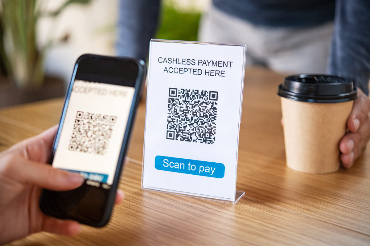 A hand holds a phone as it scans a QR on the camera app. The QR code is in a small plastic sign that indicates, "Scan to pay," while an employee holds a to-go coffee cup on the table.