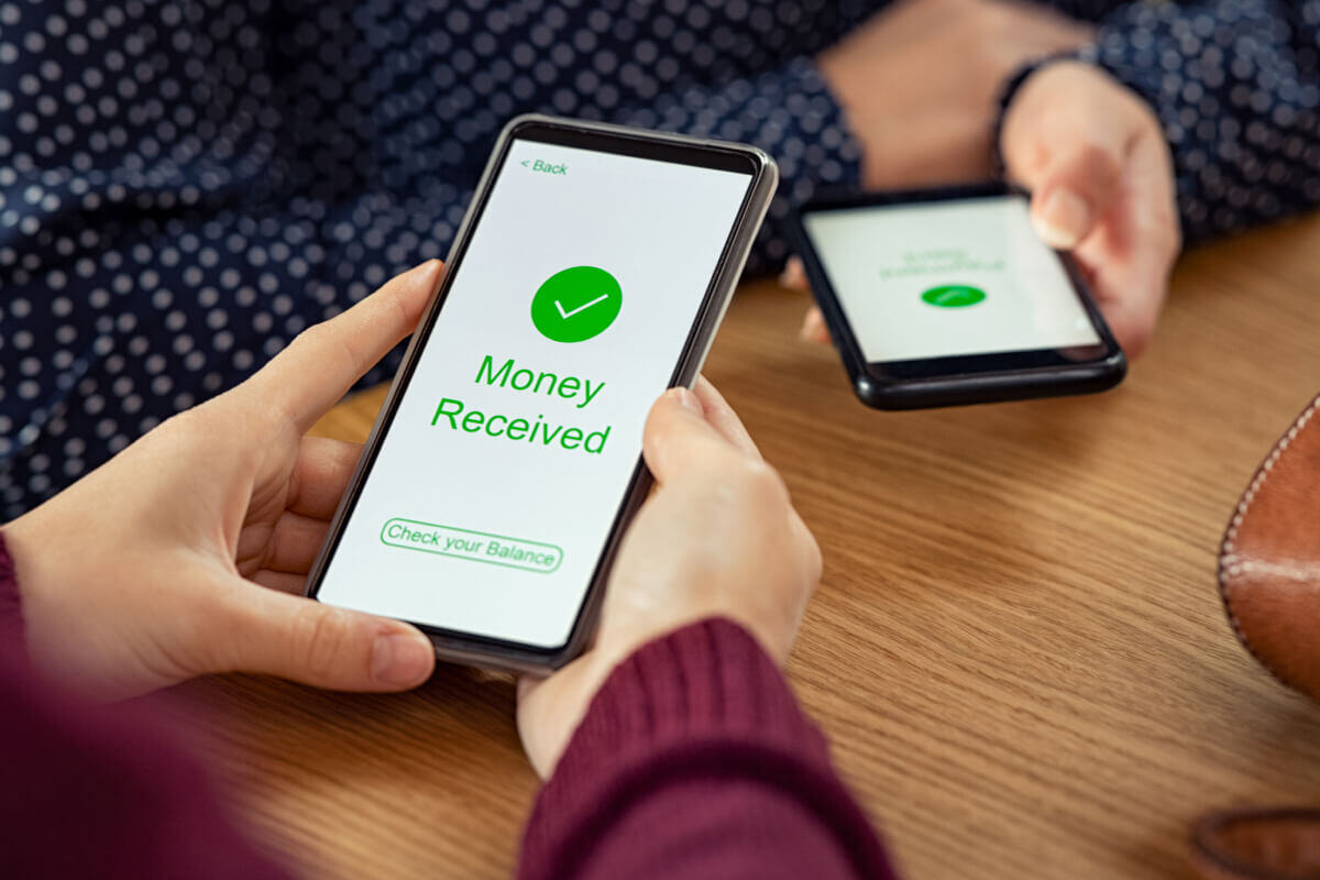 A person's hands hold a smartphone with a "Money Received" notification taking up the whole screen, showing a competed peer-to-peer transaction. In the background, another person holds their phone with the same message on it.