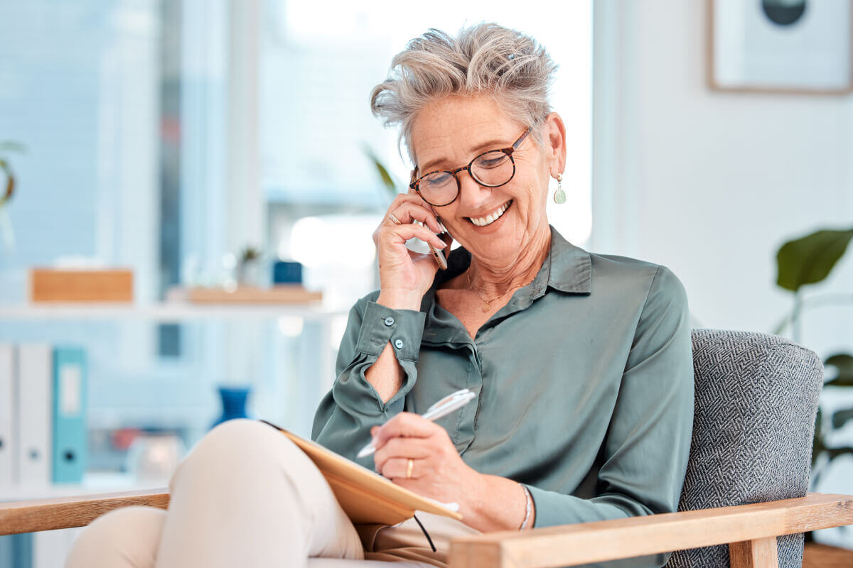 Older woman with short gray hair and brown, round glasses smiles as she makes a phone call, holding a pen and marking something off on the paper in front of her.