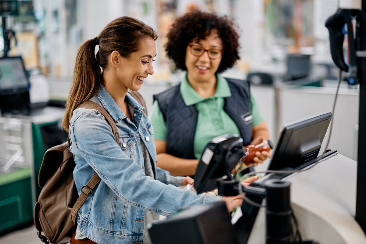 Woman in a jean jacket and wearing a brown backpack completes a purchase at a self check out machine's point of sale terminal while smiling. A worker in a green polo and navy vest smiles on in the background.