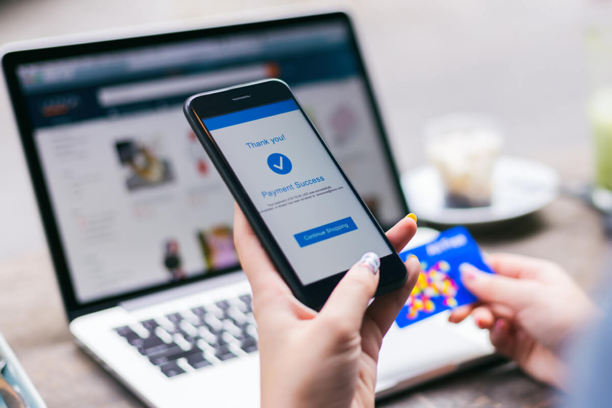 A woman's hand holds a smart phone that has a payment success page on it. Behind it, her other hand holds a credit card above an open laptop.