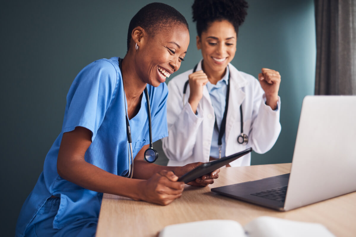 Two Black female medical professionals stand beside each other in front of an open laptop. One woman is holding a laptop, and the other woman is excitedly pumping her fists.