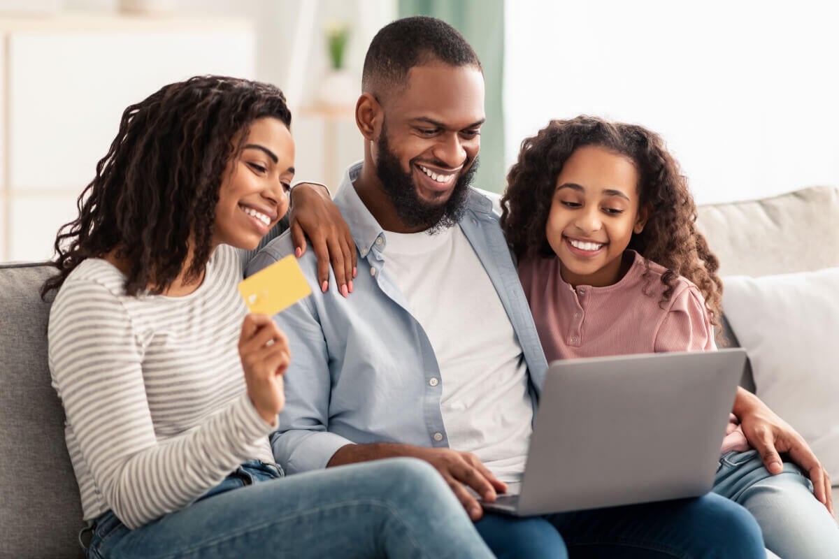 A family of three sit on the couch, smiling and looking at an open laptop. The family consists of a Black mom, dad, and child, with the mom holding a credit card while they complete a virtual payment online.