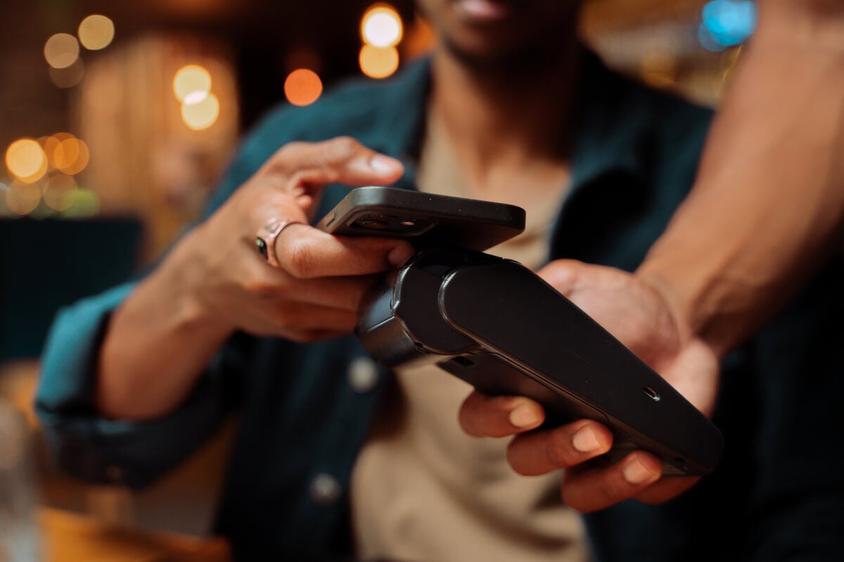 Hand wearing a silver ring holds up a black smartphone against a handheld payment terminal to complete a cashless payment. There are twinkling lights in the background as the phone holder smiles.