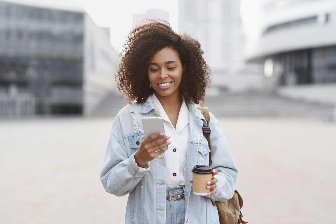 Woman on phone paying by text using FirsTech's mobile payment systems while walking and drinking coffee
